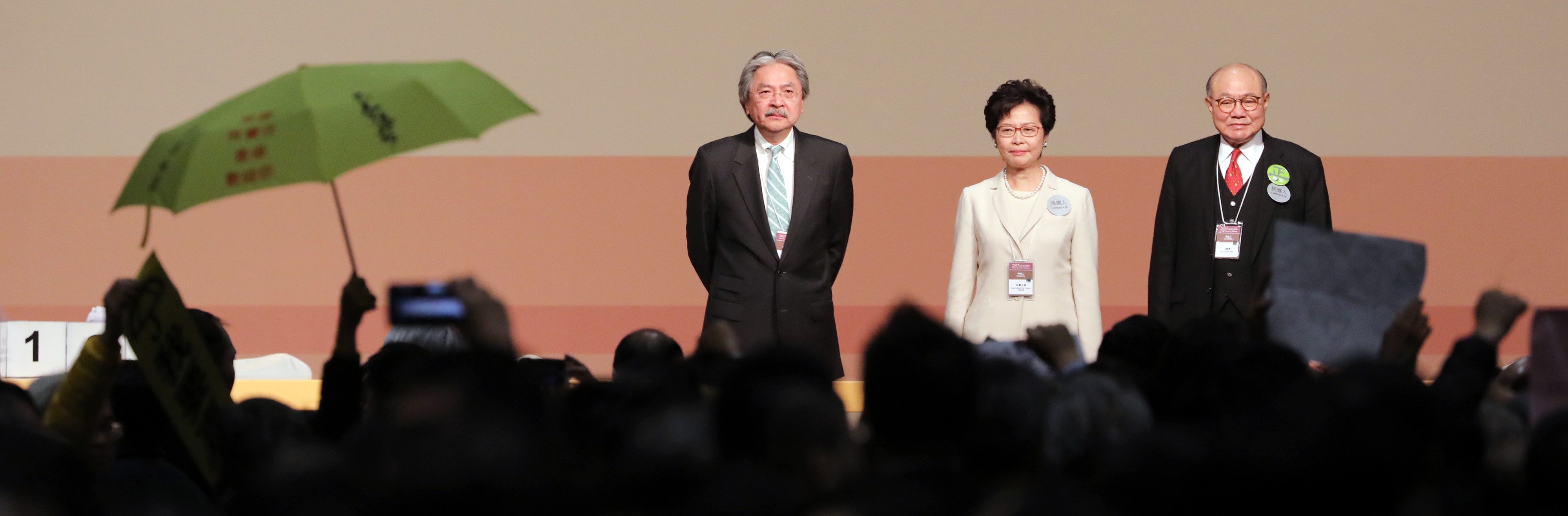 A protester raises a yellow umbrella as Chief Executive-designate Carrie Lam stands on stage with fellow candidates John Tsang and Woo Kwok-hing, March 2017 (Photo: Getty Images/Bloomberg)