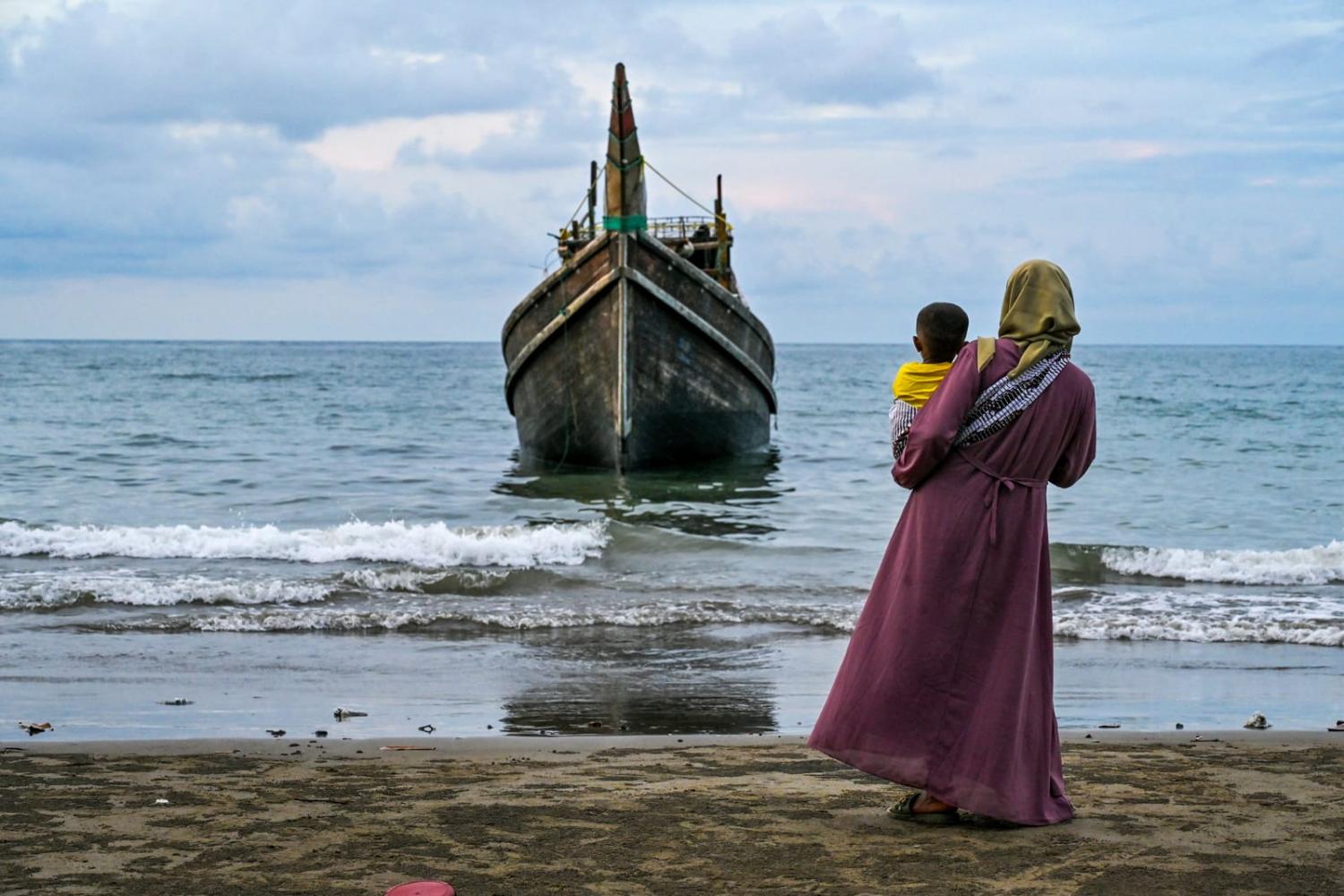 A woman with her child looks at a boat which carried an estimated 300 Rohingya refugees to Laweueng beach in Pidie district of Aceh province, 10 December 2023 (Chaideer Mahyuddin/AFP via Getty Images)